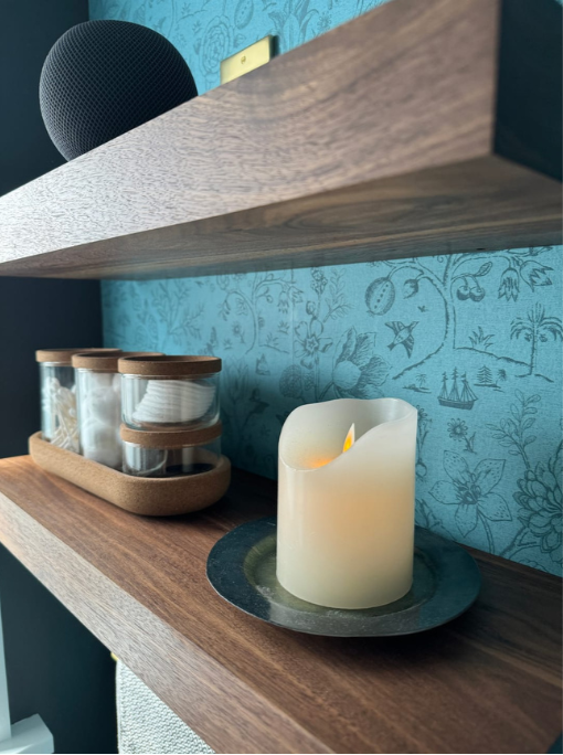 Candle on a wooden shelf with decorative jars against a blue patterned wall.